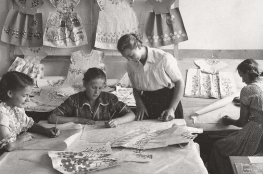 "A snapshot of a fashion design studio in black and white. An older woman instructs the young girls as they gather around a table with skirt patterns. "