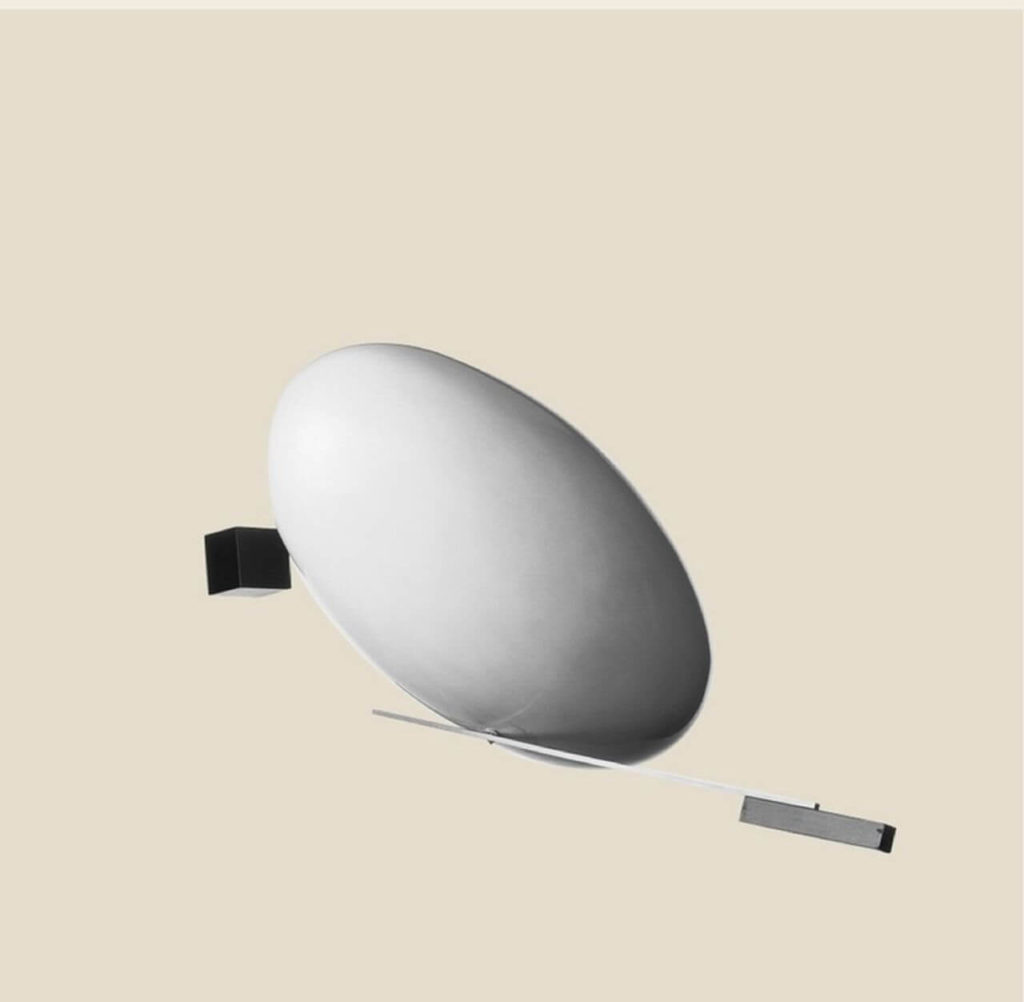 A sculpture created with a variety of materials. Though the shapes may resemble some everyday objects, it is an abstract work of art. There is an egg-like object in the center with what appears to be a long, thin blade beneath it. There is also a hanging small black square next to it.