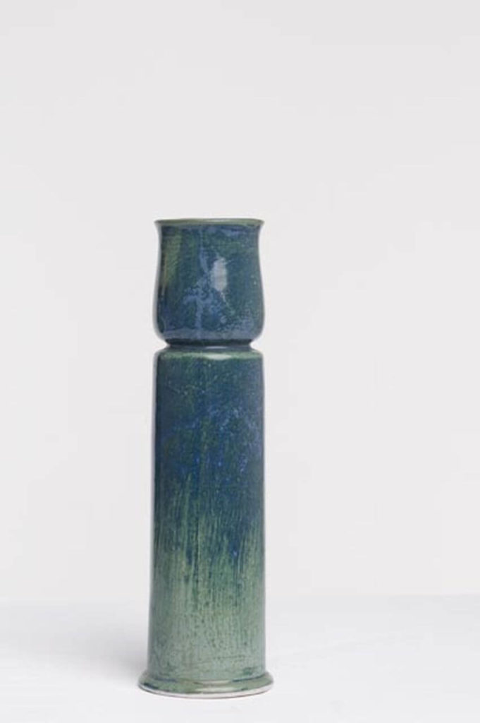 A ceramic vase of oblong shape. It is not homogeneous, with a segment clearly separated by a line at three-quarters of its height. The vase is light green, stylised to look aged, with the colour giving the impression of having been rubbed off in some places.