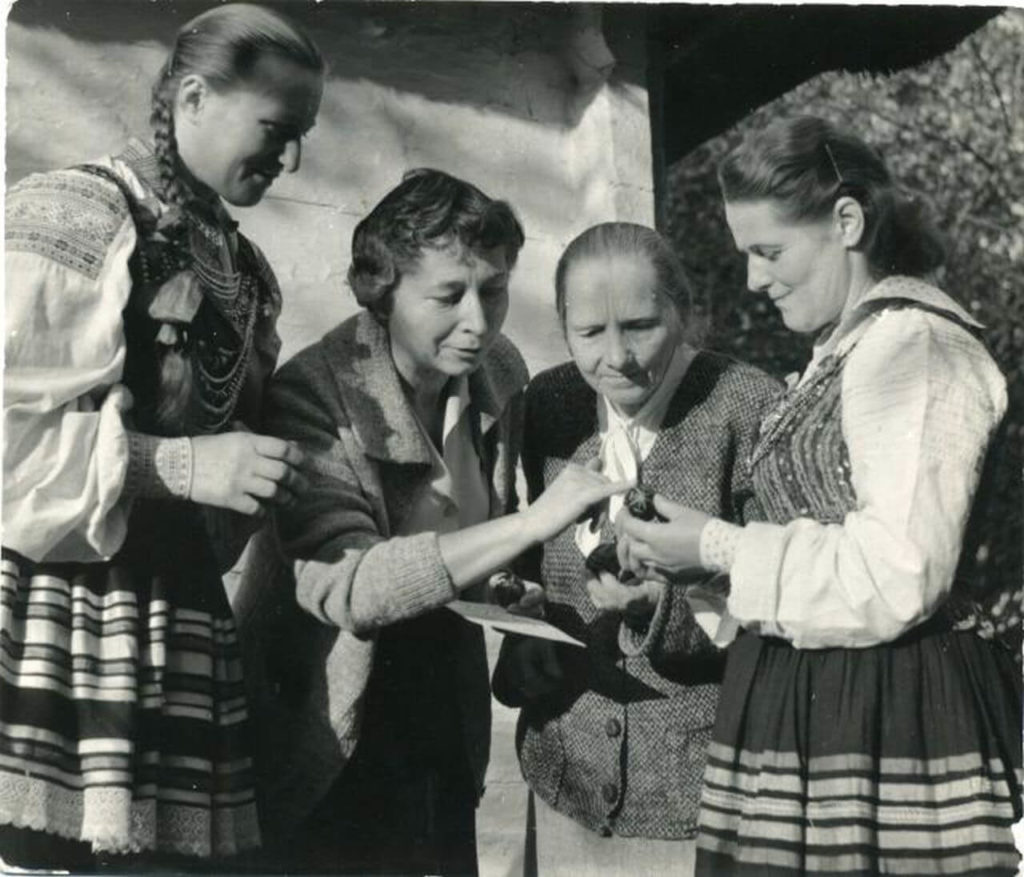 Four women of varied ages are photographed in black and white. Two of them are dressed in traditional clothing. They are all enthralled by a little object held by one of them.