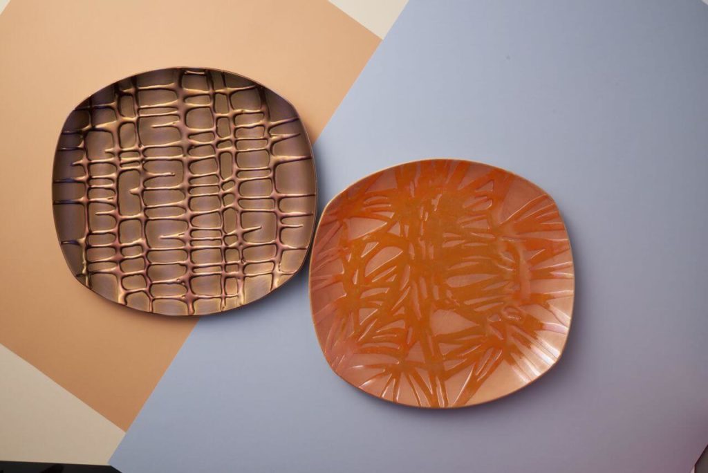 Two oval-shaped platters with shapes that remind of the CRT TV sets. The left one is brown with a semi-regular geometric pattern; the one on the right is orange, decorated with organic floral ornament. Both are coated with mirror-finish enamel.