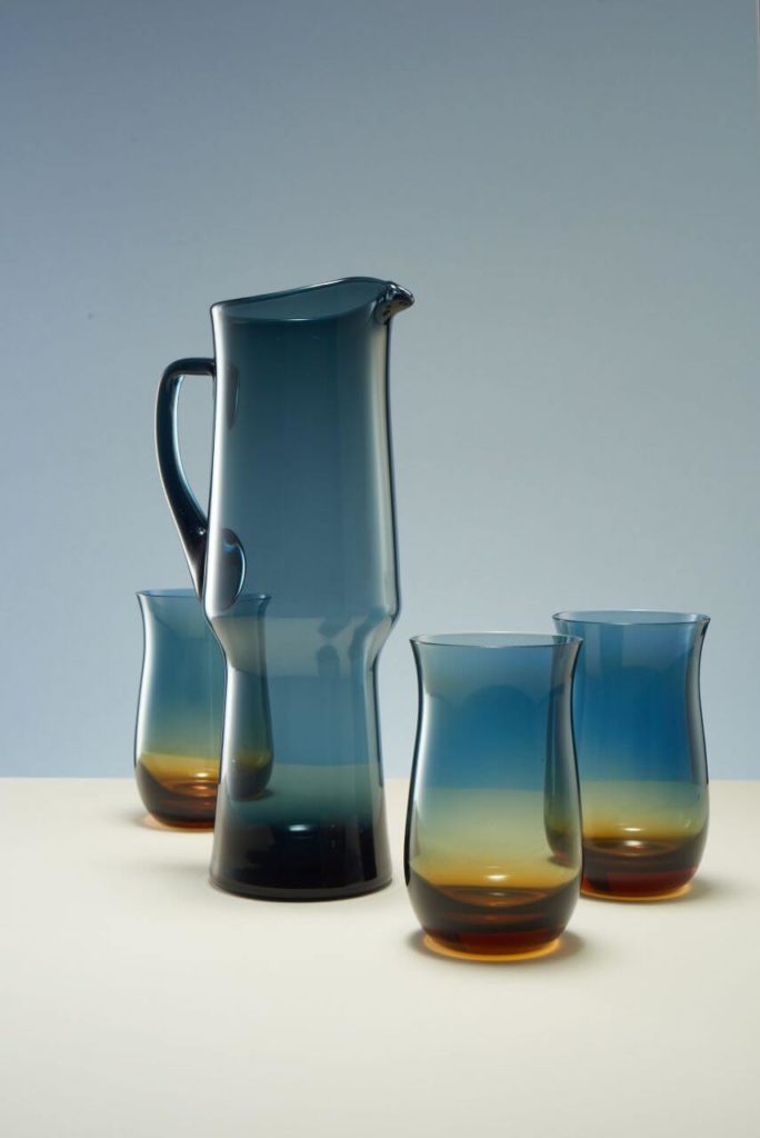 A tall jug and three glasses around it. All are made of dark glass. The upper parts are dark blue, while the bottom parts are dark brown. The jug in the lower half is narrower than in the upper half, and its handle is completely dark. The upper edges of all items are outward curved.