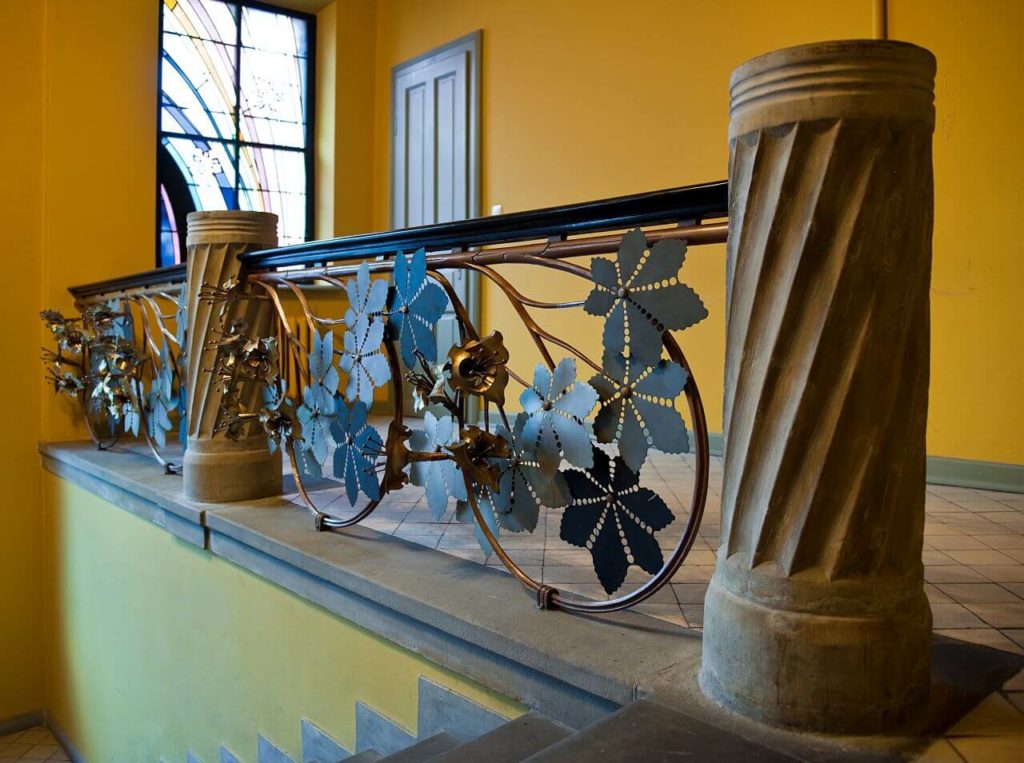 A contemporary photograph of the railing of the staircase decorated with a chestnut tree leaf motif. In the background, the stained glass window with abstract shapes is visible.
