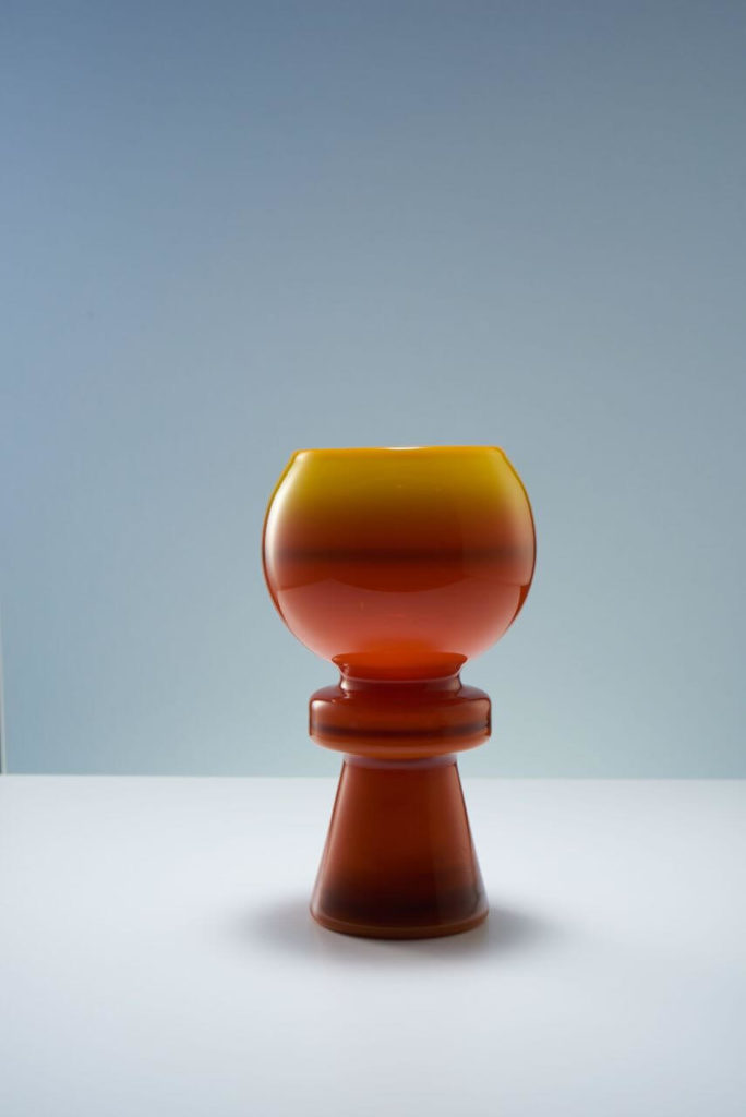 Vase made of glass. A long, tapering leg with a straight line around it serves as its foundation. It looks like a chess piece. The upper portion is shaped like a spherical bowl. The entire piece is semi-transparent, with the lower half being dark brown and the bowl being amber.
