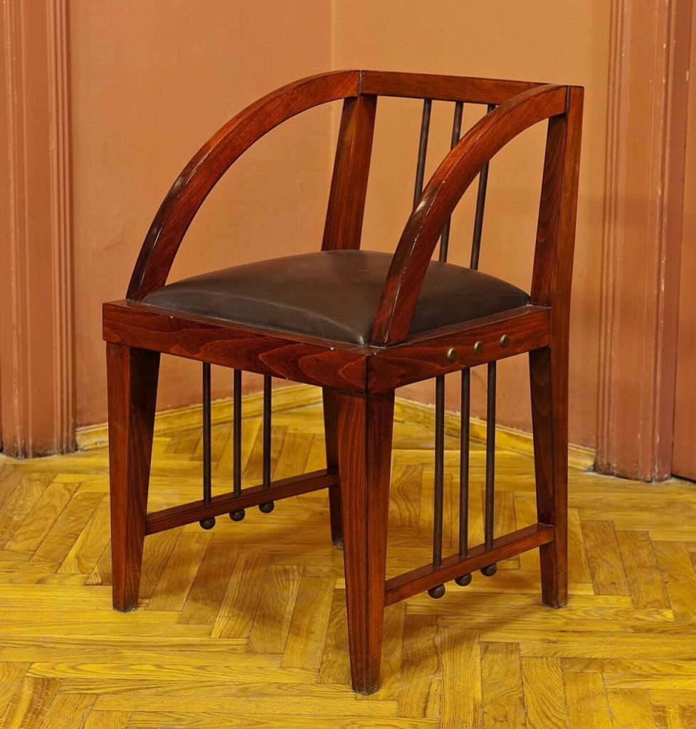A brown chair made mostly of wood. It has arc-shaped armrests that are the same height as the backrest, providing no support for the eventual sitter. The seat however is soft and prominent.