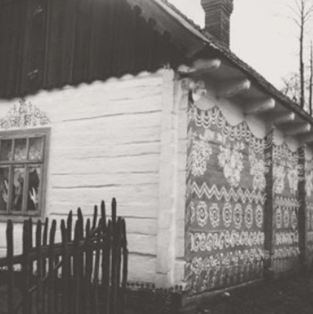 A black and white snapshot of a historic old house with folk designs on one wall. A section of a black fence can be seen on the left.