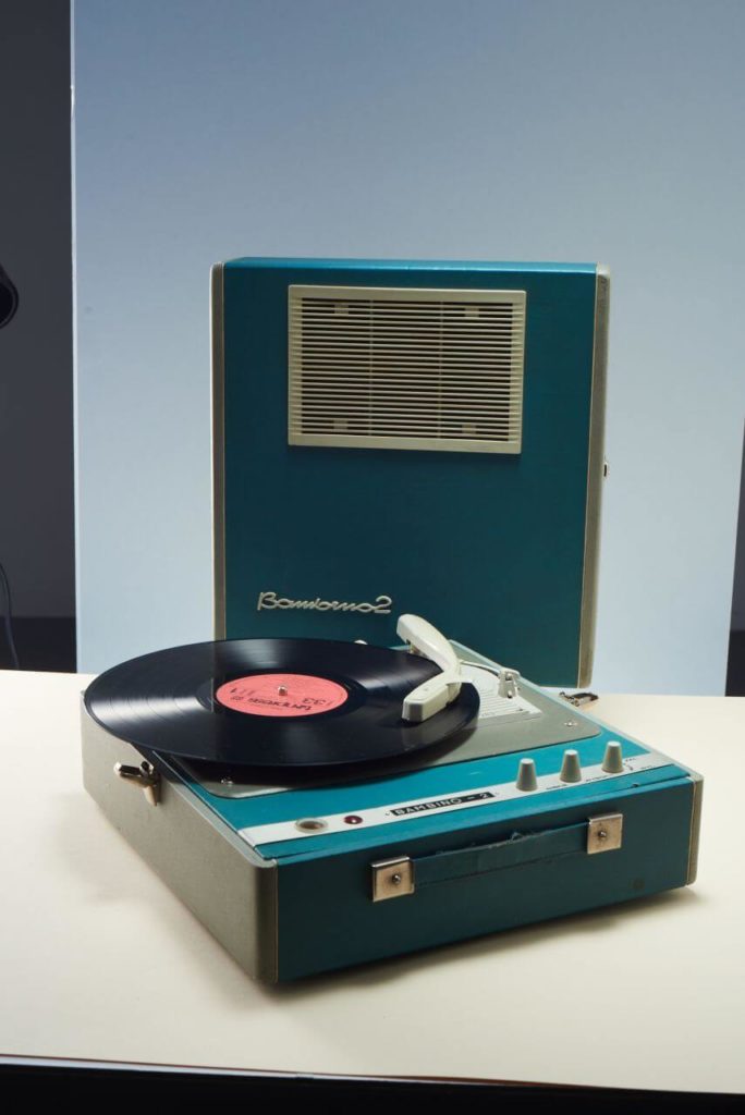 An old turntable that looks like a suitcase, playing a black vinyl record. Its removable top has a built-in speaker. It is blue and cream; its arm is white. The inscription 'Bambino - 2' is visible on the unit and under the speaker.