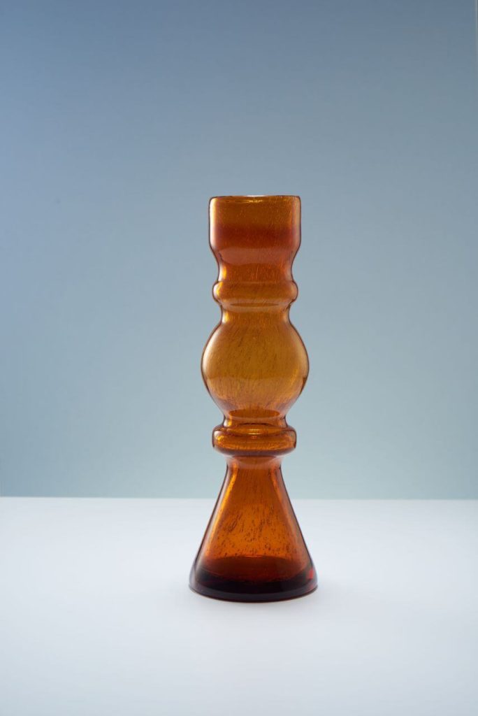 A semi-transparent candle holder. Its round base is dark-brown, while upper parts are brighter: from light-brown to amber. The shape resembles a laboratory flask. Its foot is triangular, the central part is spherical, and the upper part is straight.