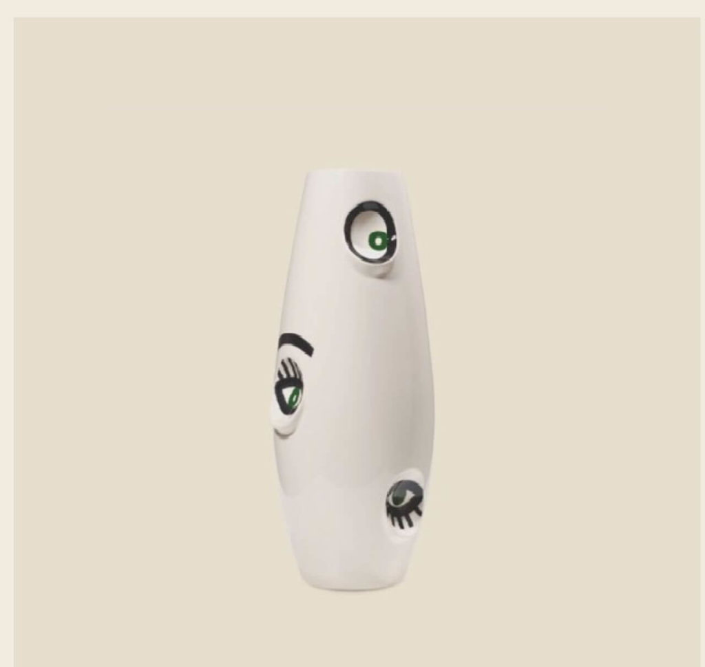 A white ceramic spindle-shaped vase with painted human eyes. The eyes have black borders and green irises.