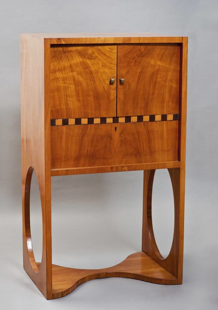 A wooden, light brown dressing table. The construction consists of two shelves. The upper one can be opened using double doors. The lower one has a keyhole. There is a thin frieze in between with a pattern of black and yellow squares. The base of the table is a unified structure. The two O-shaped legs are joined by an hourglass-shaped board lying on the floor..